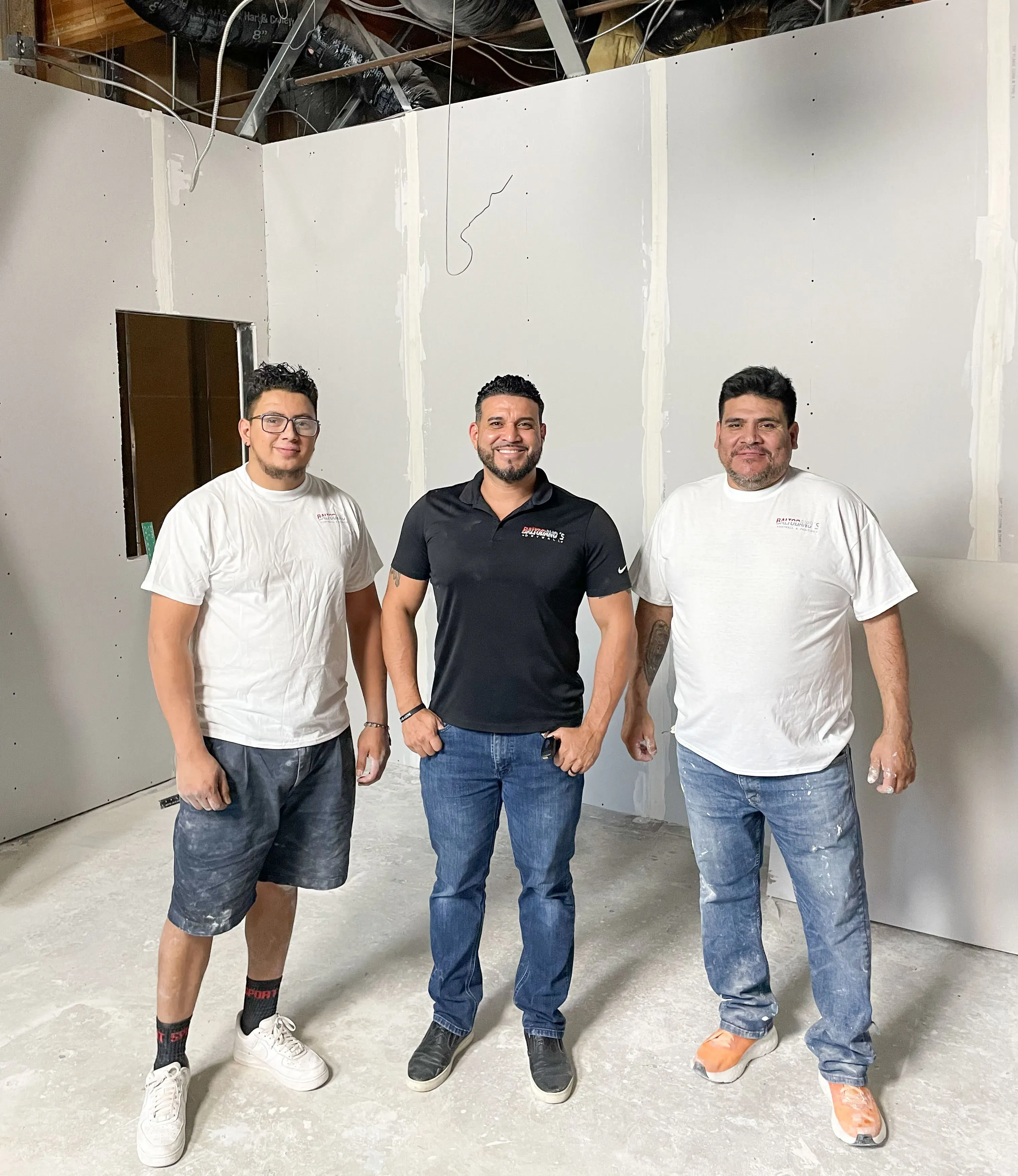 "Three individuals standing in front of a drywall installation, wearing casual work attire, representing Baltodano's Drywall and Painting in Oakley."